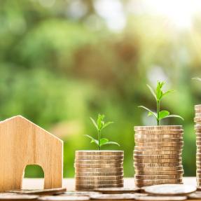 a miniature wooden cutout of a house next to increasingly tall stacks of coins with plant sprouts growing out of the top of the stacks